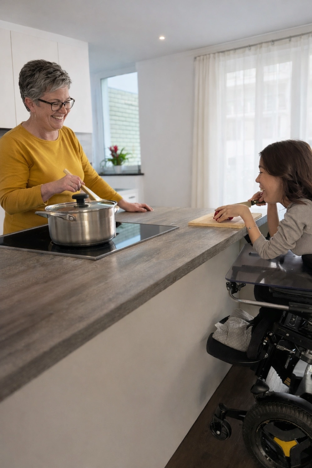 Hadzsi GbR - Assistenzdienstleistungen - Eine behinderte Frau sitzt in einem Elektrorollstuhl und schneidet Gemüse, um einer Frau in der Küche zu helfen.